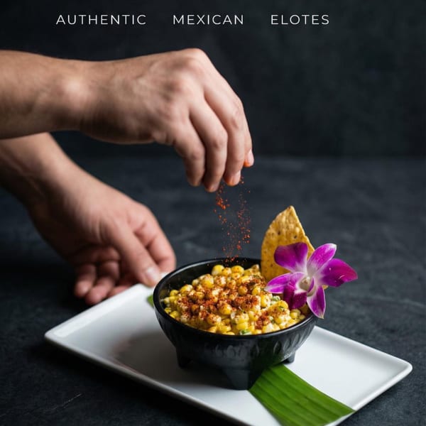 Hands sprinkling chili powder over a bowl of elotes, garnished with a tortilla chip and orchid.